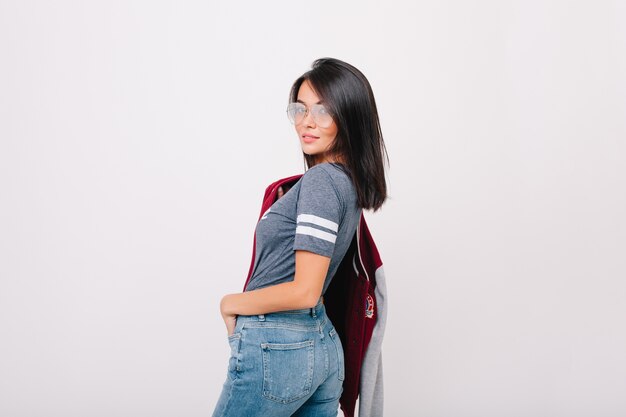 Refined girl with short black hair looking over shoulder, holding bomber in other hand. Pretty lady with trendy haircut having fun and posing with pleasure in new glasses.