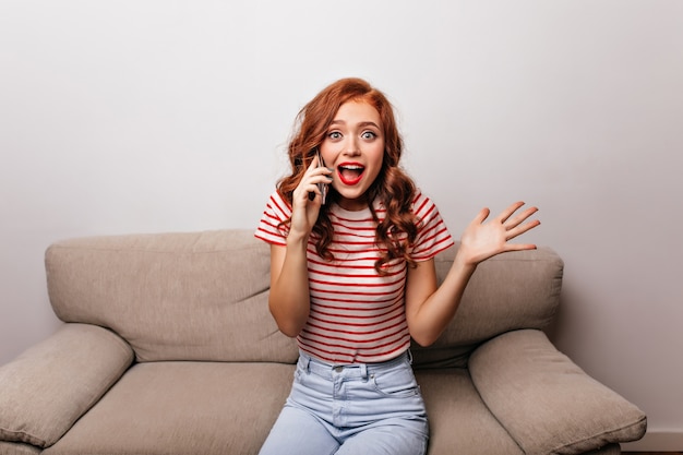 Refined lady with curly hairstyle talking on phone. Pretty ginger girl sitting on sofa and calling someone.