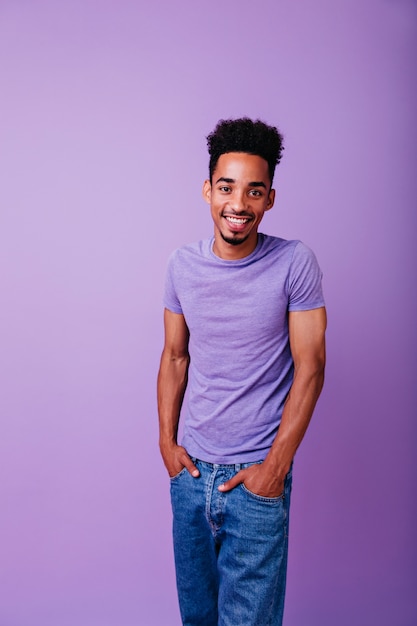 Refined male model with curly hair standing with happy face expression. Indoor shot of glad young man holding hands in pockets.