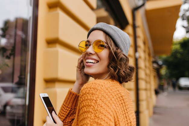 Refined short-haired woman in sunglasses looking over her shoulder while posing on the street