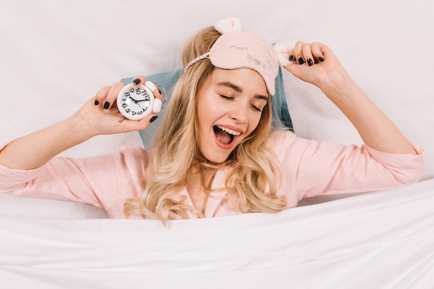 Refined young woman in pink sleep mask posing with clock