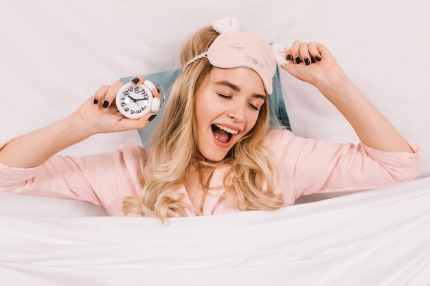 Утонченная молодая женщина в розовой маске для сна позирует с часами
