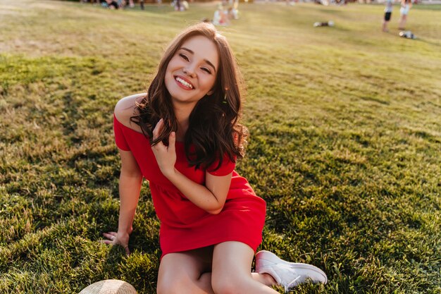 Утонченная молодая женщина в красном платье сидит на траве. Веселая улыбающаяся девушка с темными волосами позирует на лужайке.