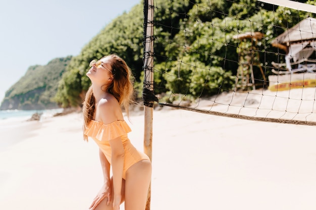 Raffinata giovane donna indossa costume da bagno alla moda in posa con piacere vicino al set di pallavolo. colpo all'aperto di incredibile ragazza dai capelli castani che gode del tempo libero attivo in spiaggia.