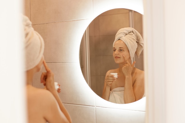 鏡で化粧クリームを顔にこすりつけ、バスルームで顔の皮膚に保湿剤を塗り、白いタオルに包まれ、美容処置をしている女性の反射。