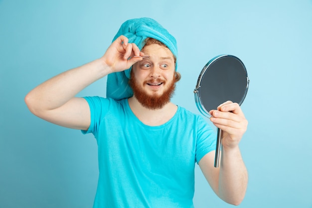 Reflection. portrait of young caucasian man in his beauty day and skin care routine. male model with red hair making up his eyebrow using mirror. body and face care, natural, male beauty concept.