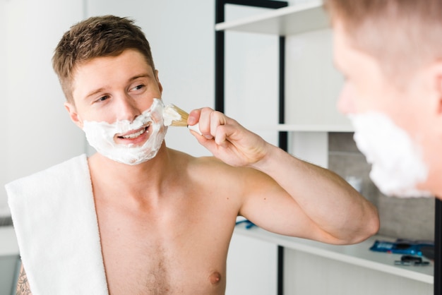 Reflection of shirtless young man with white towel over his shoulder shaving with brush