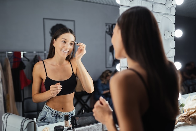 Отражение молодой красивой женщины, применяя ее макияж, глядя в зеркало