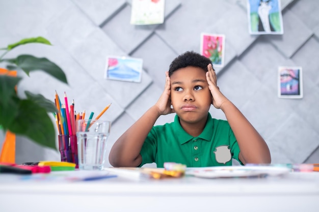 Reflections. Dark-skinned boy of primary school age touching head with hands looking pensively up and aside sitting at table with drawing and colored pencils in room