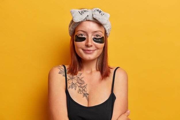 Refreshed female dressed in black t shirt, soft headband, applies collagen patches for reducing puffiness and dark circles, cares of skin around eyes. Facial treatment concept. Morning routine