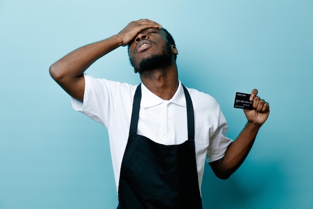 Regret holding card putting hand on forehead young african american barber in uniform isolated on blue background