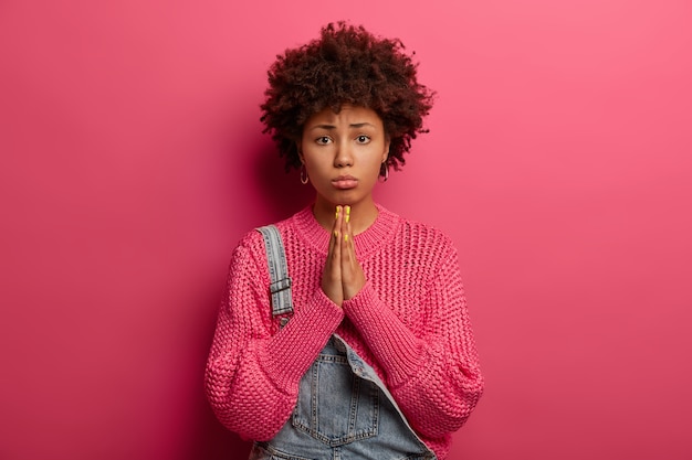 Free photo regretful dark skinned african american woman beggs for forgiveness, feels guilty, purses lower lip, wears knitted sweater, isolated on pink wall, beseeches parents for money, looks innocent
