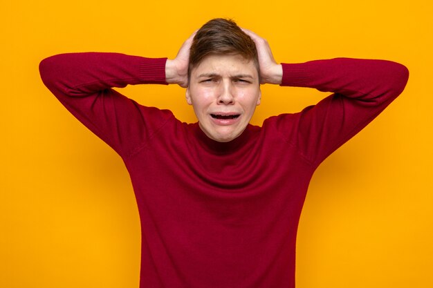 Regretted grabbed head young handsome guy wearing red sweater isolated on orange wall