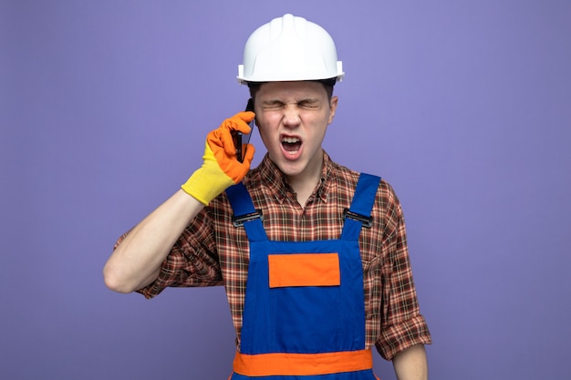 Regretted with closed eyes young male builder wearing uniform with gloves speaks on phone 