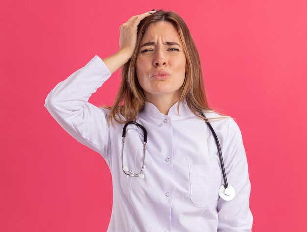 Regretted young female doctor wearing medical robe with stethoscope putting hand on head isolated on pink wall