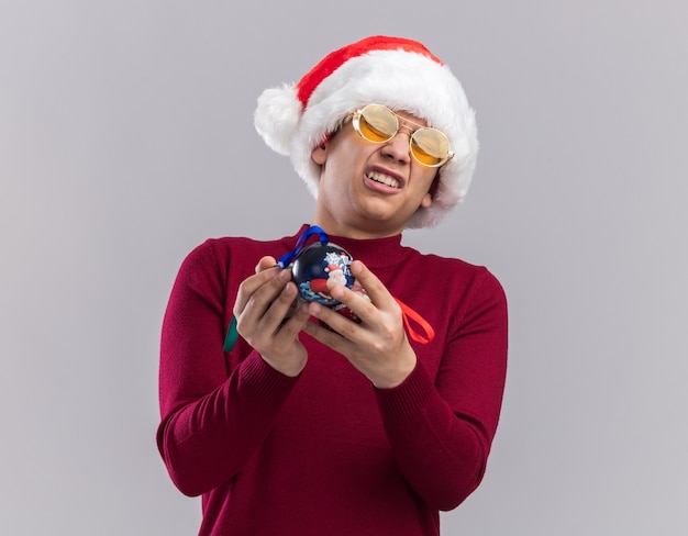 Regretted young guy wearing christmas hat with glasses holding christmas tree balls isolated on white background