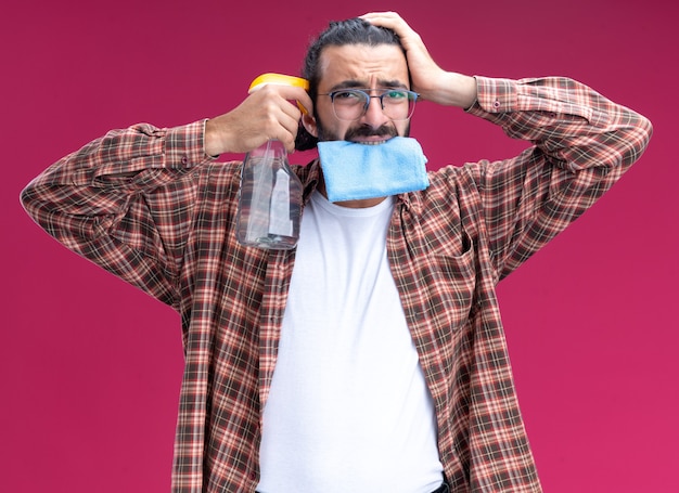 Regretted young handsome cleaning guy wearing t-shirt holding rag in mouth and putting spray bottle on temple putting hand on head isolated on pink wall