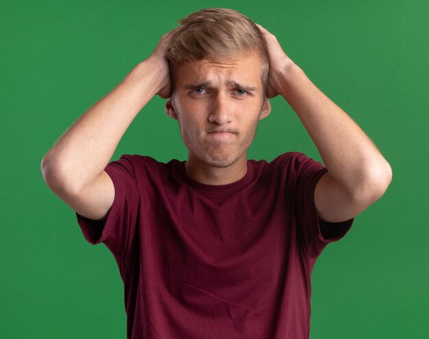 Giovane ragazzo bello dispiaciuto che indossa la camicia rossa che mette le mani dietro la testa isolata sulla parete verde