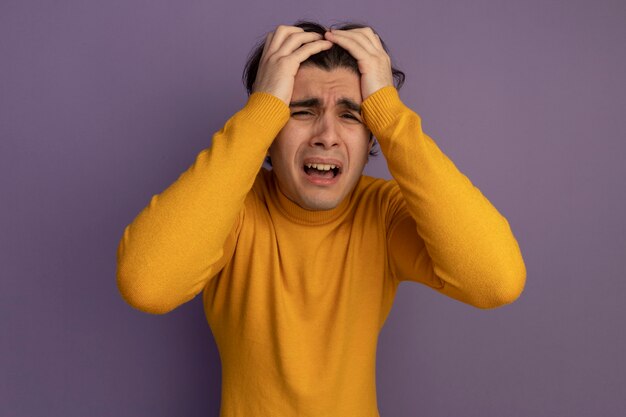 黄色のタートルネックのセーターを着ている後悔した若いハンサムな男は、紫色の壁に分離された頭をつかんだ