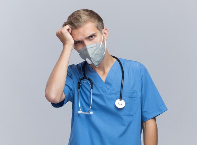 Жалко молодой мужчина-врач в униформе врача со стетоскопом и медицинской маской, положив руку на голову, изолированную на белой стене