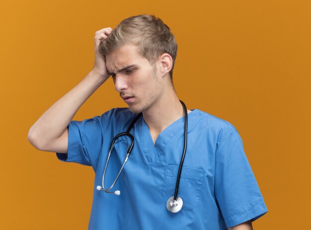 Regretted young male doctor wearing doctor uniform with stethoscope scratching head isolated on orange wall