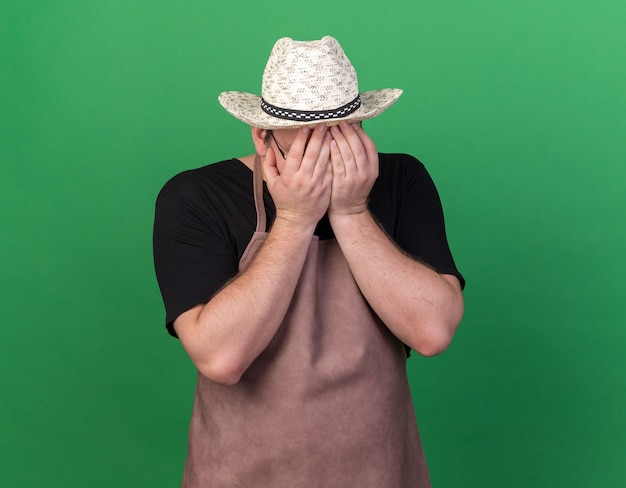 Regretted young male gardener wearing gardening hat covered face with hands isolated on green wall