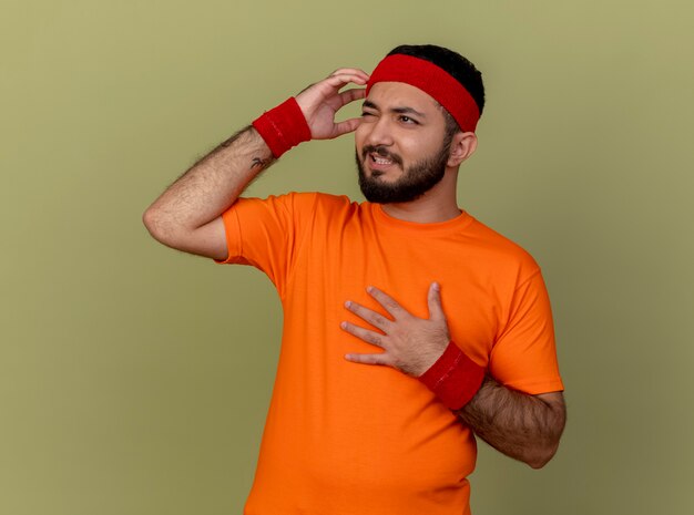 Regretted young sporty man looking at side wearing headband and wristband putting hands on chest and temple isolated on olive green background