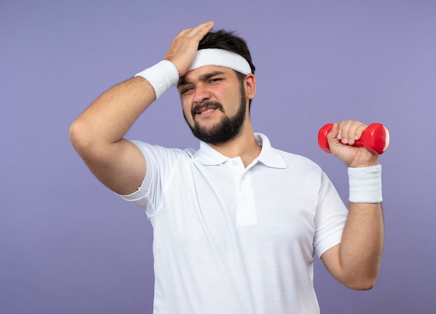 Бесплатное фото Сожалеющий молодой спортивный мужчина с повязкой на голову и браслетом, держащим гантель, положив руку на лоб