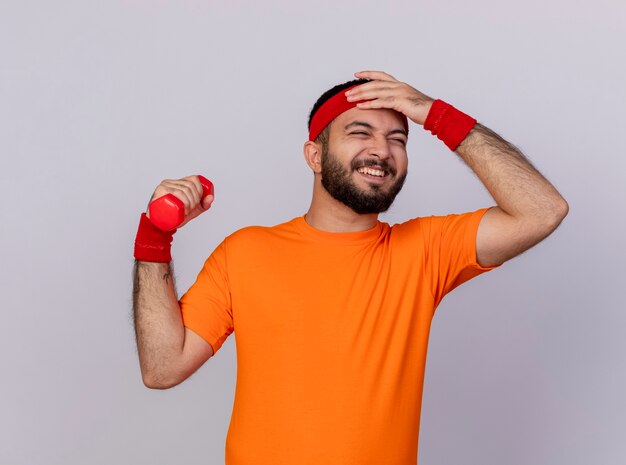 Regretted young sporty man wearing headband and wristband raising dumbbell putting hand