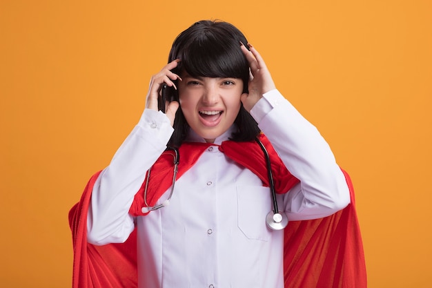 Free photo regretted young superhero girl wearing stethoscope with medical robe and cloak holding phone and grabbed head