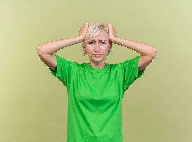 Regretting middle-aged blonde slavic woman looking at camera holding head isolated on olive green background