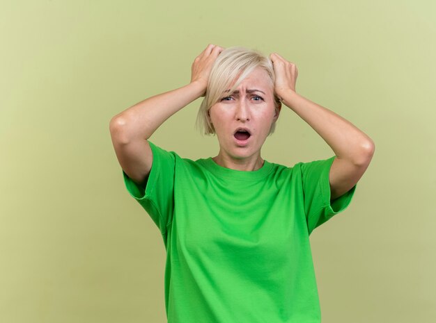 Regretting middle-aged blonde slavic woman looking at camera putting hands on head isolated on olive green background with copy space