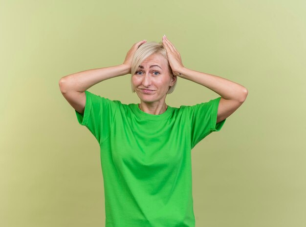 Deplorando la donna slava bionda di mezza età che guarda davanti mantenendo le mani sulla testa isolata sulla parete verde oliva