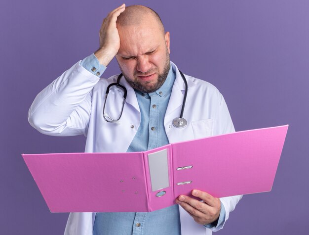 Regretting middle-aged male doctor wearing medical robe and stethoscope keeping hand on head holding open folder looking at it isolated on purple wall