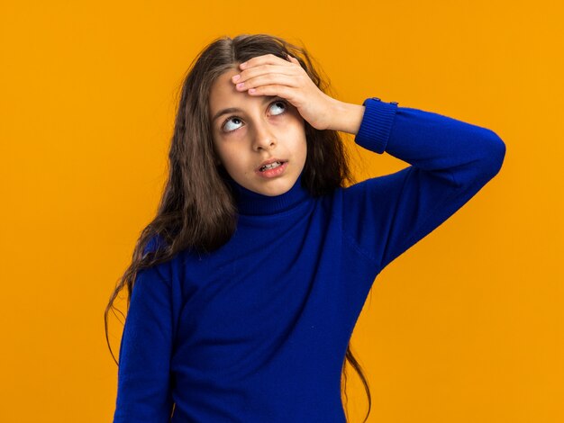 Regretting teenage girl keeping hand on forehead looking up isolated on orange wall