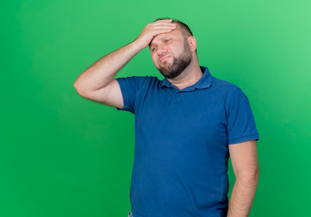 Regretting and tired adult slavic man looking straight putting hand on head isolated on green wall with copy space