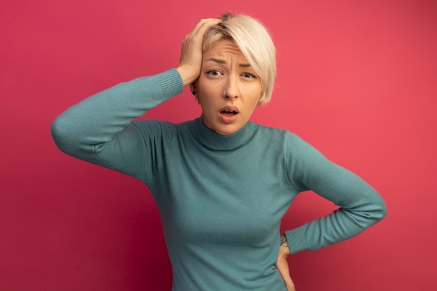 Free photo regretting young blonde girl putting hand on head and keeping another one on waist looking  isolated on pink wall