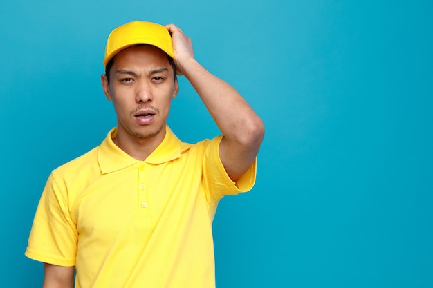 Regretting young delivery man wearing uniform and cap keeping hand on head 