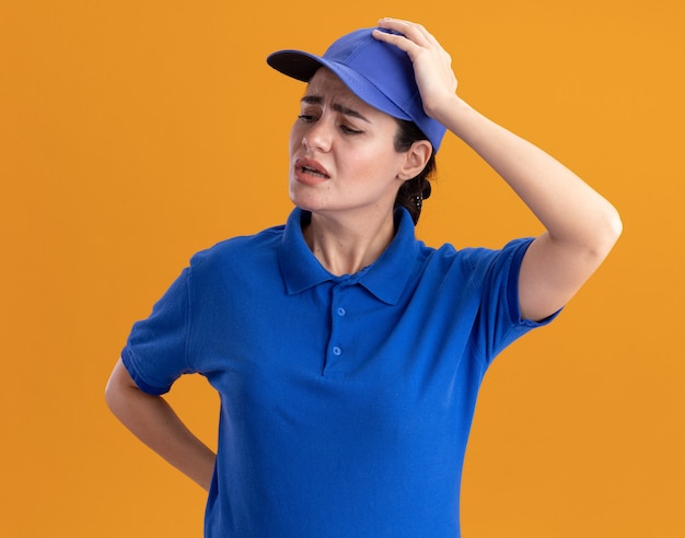 Regretting young delivery woman in uniform and cap keeping hand behind back and on head looking at side 