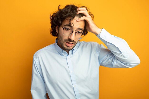 Regretting young handsome caucasian man wearing glasses putting hand on head looking down isolated on orange background