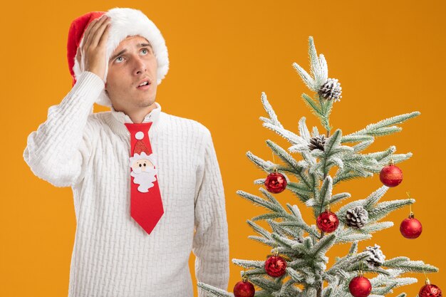 크리스마스 모자와 산타 클로스 넥타이를 착용하고 후회하는 젊은 잘 생긴 남자가 장식 된 크리스마스 트리 근처에 서서 머리에 손을 넣어 오렌지 벽에 고립 된 찾고