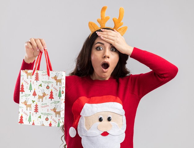 Сожалеющая молодая красивая девушка в повязке на голову из оленьих рогов и свитере санта-клауса, держащая рождественский подарочный пакет, глядя, держа руку на лбу