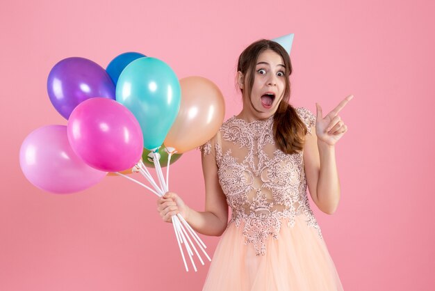 rejoiced girl with party cap holding balloons on pink