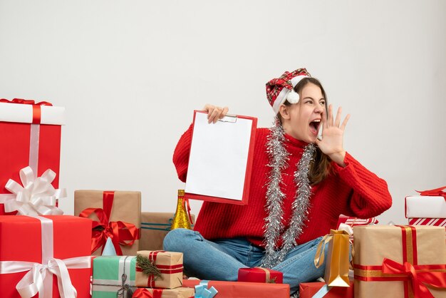 rejoiced girl with santa hat holding files and shouting sitting around presents on white