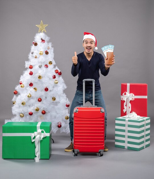 rejoiced man with red suitcase showing his travel tickets and making thumb up sign on grey