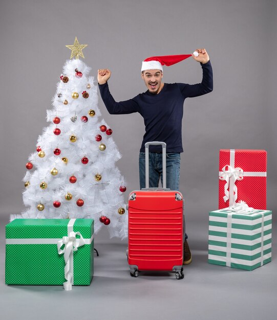 흰색 크리스마스 트리 근처에 서있는 그의 산타 모자를 들고 기뻐 젊은 남자와 회색 선물