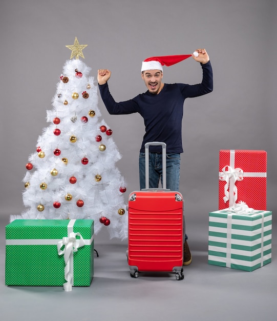 rejoiced young man holding his santa hat standing near white xmas tree and presents on grey