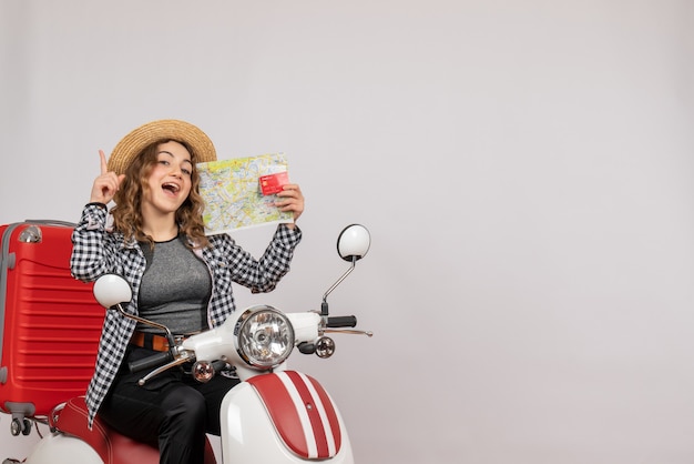 rejoiced young woman on moped holding card and map on grey