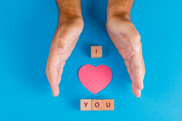Relationship concept with wooden cubes on blue table flat lay. hands protecting paper cut heart.