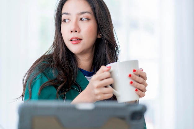 Расслабьтесь после работы, азиатская женщина-врач рука держит чашку горячего кофе, случайный отдых после тяжелой работы телемедицины в клинике в то время как носить униформу